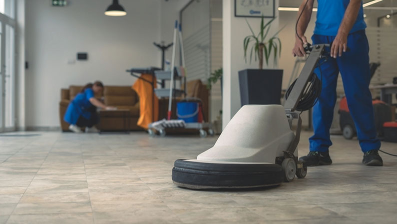 Worker polishing hard floor with high speed polishing machine while other cleaner cleans rhe table in the background