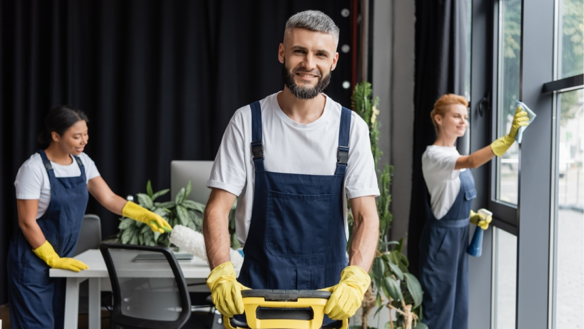 happy man with floor cleaning machine looking at camera near multiethnic cleaning offices