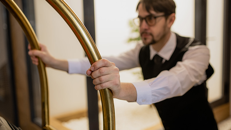 Hand of young porter holding by handles while pushing cart and picking up luggage of hotel guests