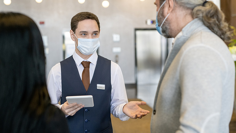 Young male receptionist in protective mask and formalwear using touchpad while talking to mature businessman in the lounge