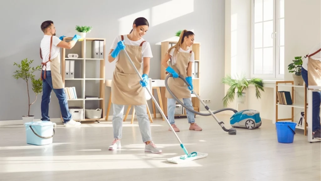 Happy smiling young cleaners men and women in uniform wiping desk, mopping floor and vacuuming. Cheerful team of professional janitors washing office workplace. Cleaning service concept.