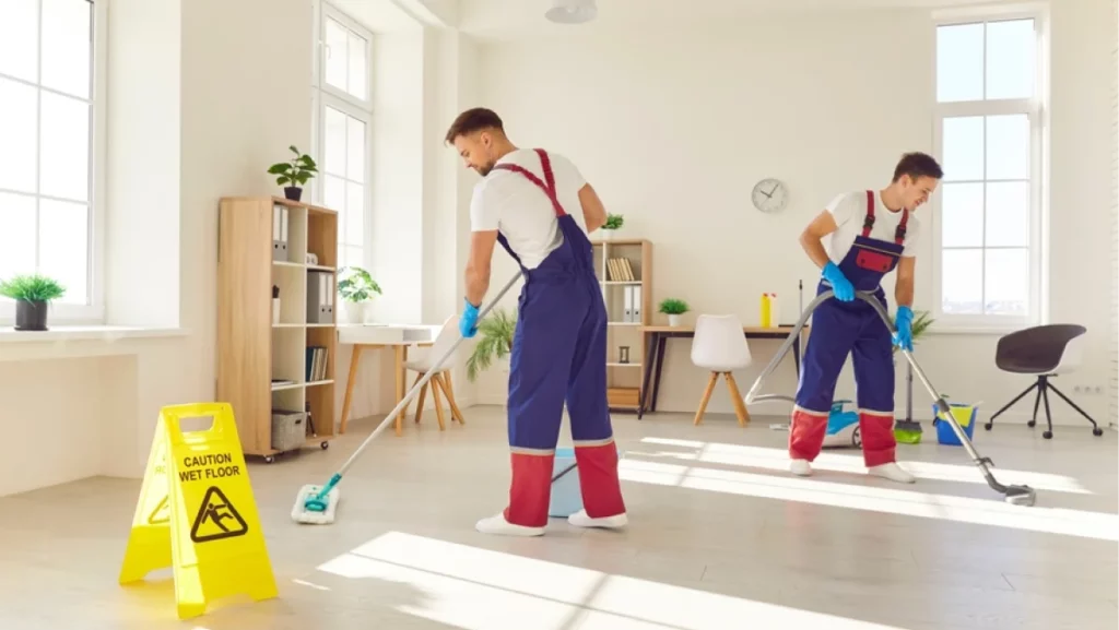 Efficient cleaning service in action. Two professional cleaners in uniform diligently clean floor in modern apartment. Male employees of cleaning company use signs with inscription caution wet floor.