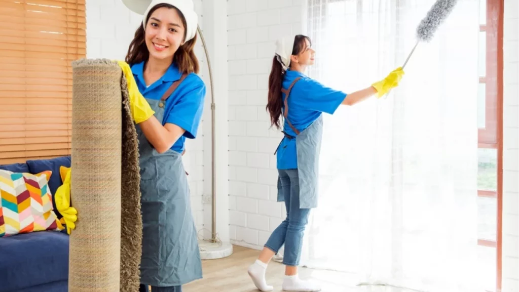 Two Asian young professional cleaning service women worker team working in the house. Girl housekeeper holding rolled carpet before cleaning wooden floor. Cleaning service. Photo with copy space